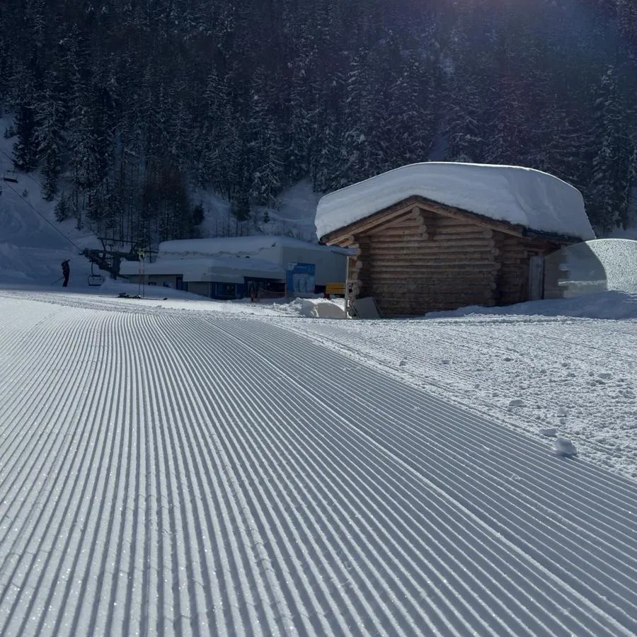 Świeżo przygotowana trasa narciarska z widocznym drewnianym budynkiem pokrytym śniegiem i wyciągiem krzesełkowym w tle.