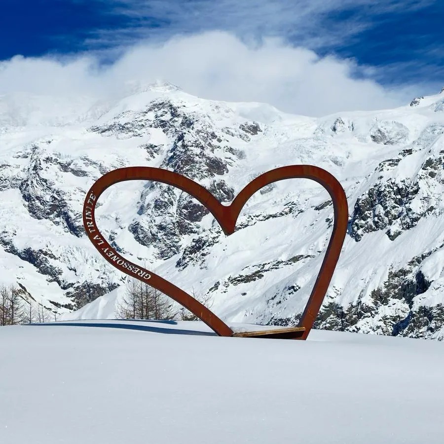 Metalowa konstrukcja w kształcie serca z napisem 'GRESSONEY LA TRINITE' na tle ośnieżonych gór i błękitnego nieba z chmurami.