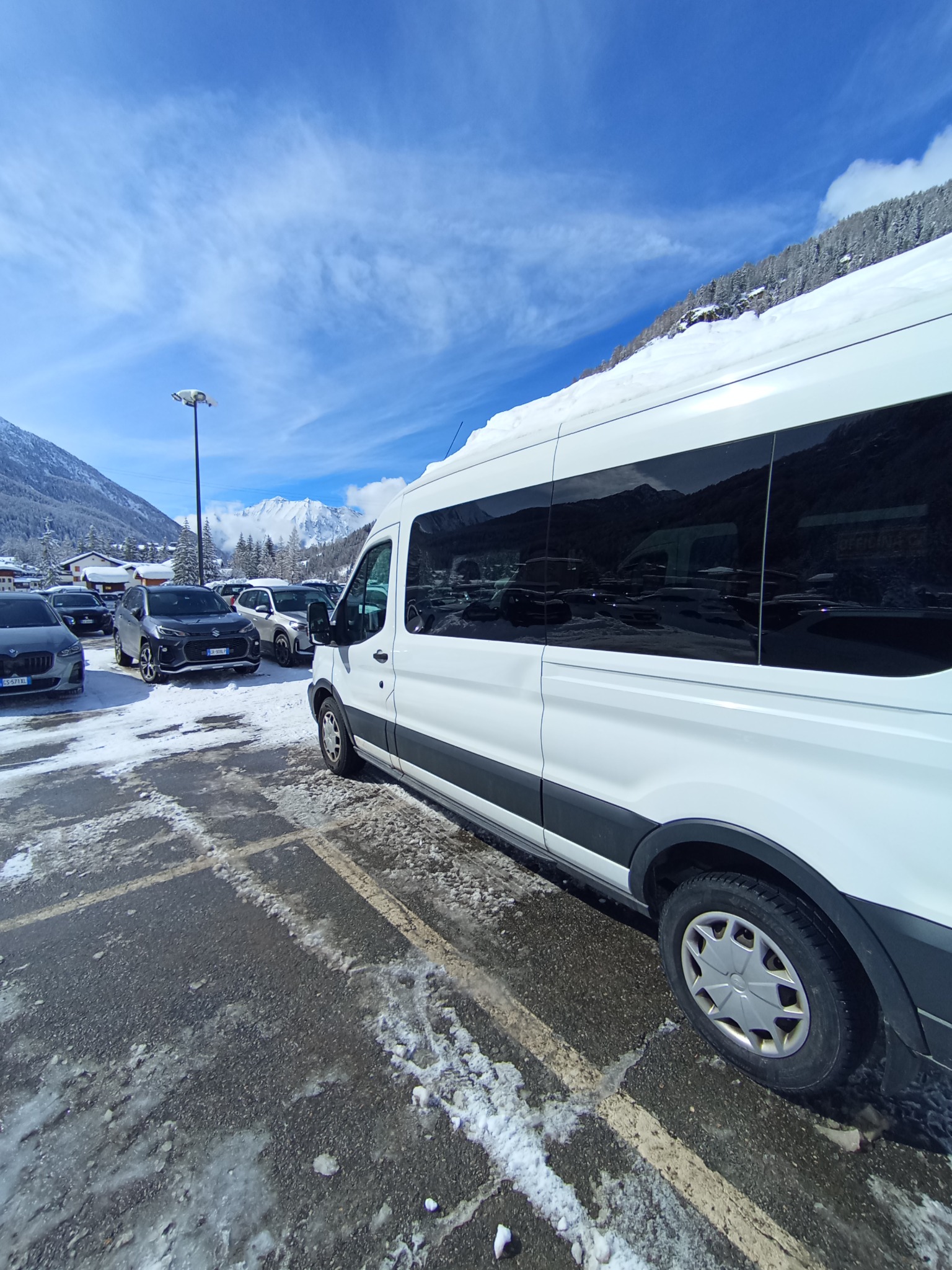 Biały bus na parkingu w zimowej scenerii, z zaśnieżonymi górami w tle, widok z boku.