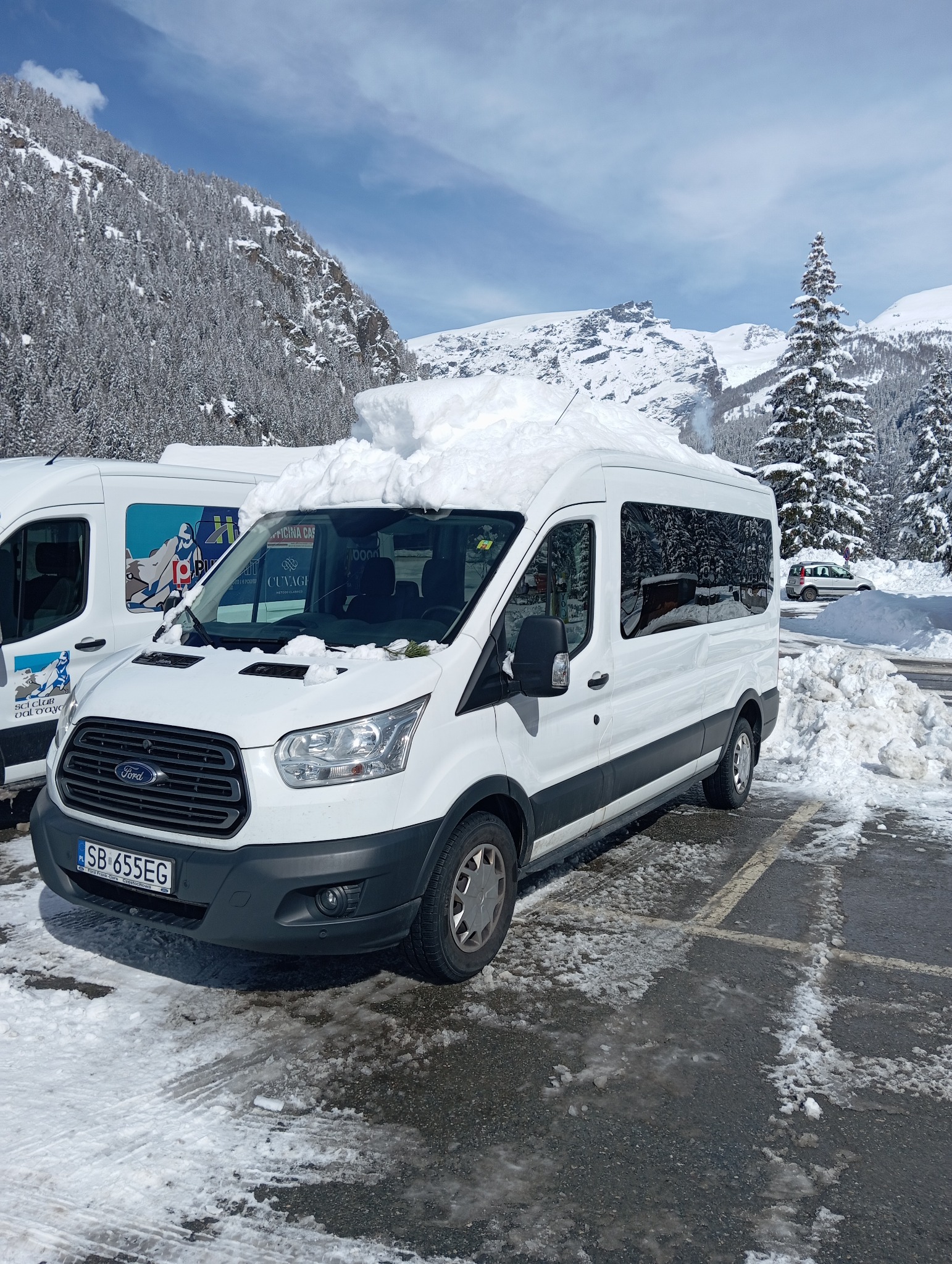 Biały bus Ford Transit zaparkowany na ośnieżonym parkingu, z górą śniegu na dachu, w tle ośnieżone góry i drzewa.