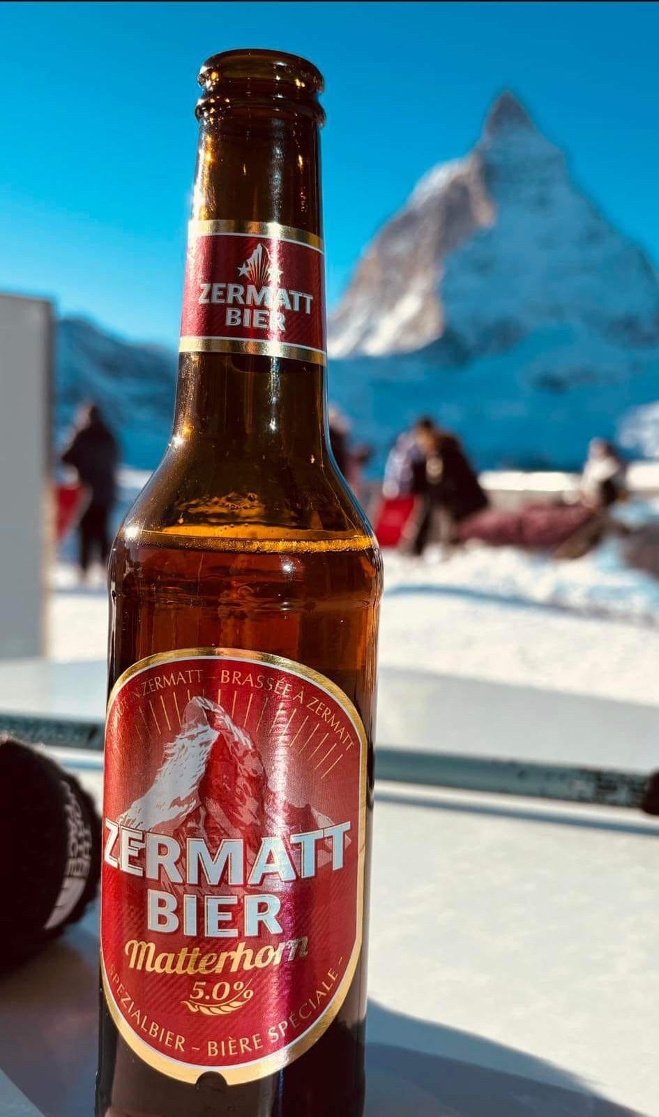 Butelka piwa Zermatt Bier na tle ośnieżonego Matterhornu i narciarzy na stoku.