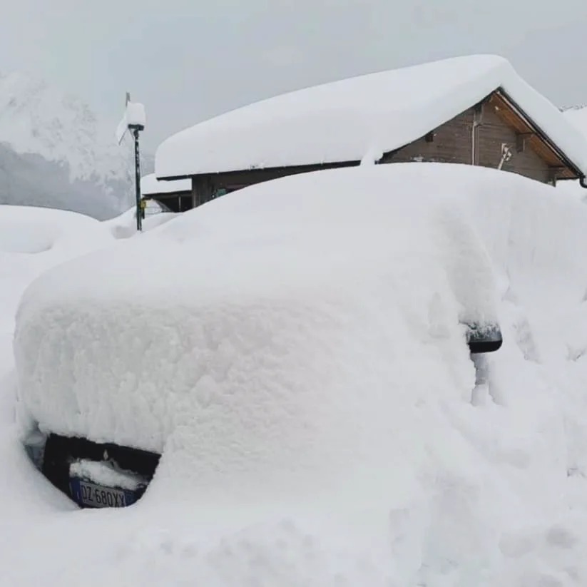 Samochód osobowy przysypany grubą warstwą śniegu, widoczny fragment tablicy rejestracyjnej i zarys budynku w tle.