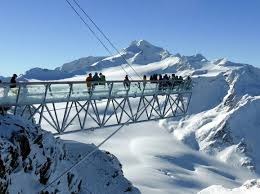 Grupa osób na przeszklonym tarasie widokowym, konstrukcja stalowa, w tle ośnieżone szczyty górskie i błękitne niebo.