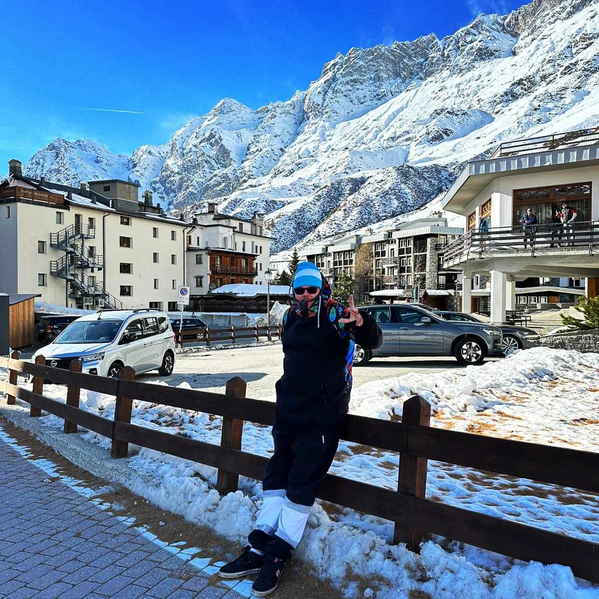 Osoba w zimowym stroju opiera się o drewniane ogrodzenie na tle ośnieżonych gór i zabudowań miejskich, z zaparkowanymi samochodami w tle.