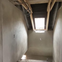 FAMILY-TYNK Dominik Maniecki - Wnętrze poddasza w trakcie prac wykończeniowych, widoczne surowe tynki na ścianach, drewniana konstrukcja dachu z oknem dachowym, oraz kable elektryczne zwisające z sufitu.