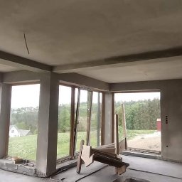FAMILY-TYNK Dominik Maniecki - Wnętrze w stanie surowym z widokiem na krajobraz górski przez duże okna, ściany i sufit pokryte świeżym tynkiem, widoczne narzędzia i resztki materiałów budowlanych.