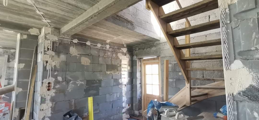 Surowy stan wnętrza budynku w trakcie remontu: ściany z szarego betonu komórkowego z widocznymi instalacjami elektrycznymi, częściowo pokryte zaprawą, drewniane schody na piętro, surowy betonowy...