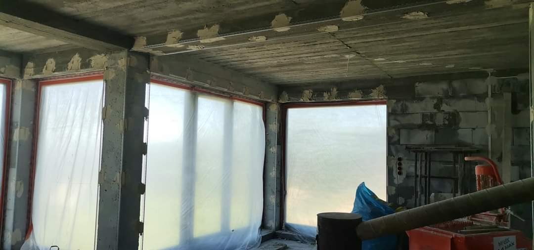 Surowy stan wnętrza z widocznymi przygotowaniami do prac wykończeniowych: okna zabezpieczone folią, ściany z betonu komórkowego z wstępnie nałożonym materiałem, widoczne szyny montażowe pod sufitem.