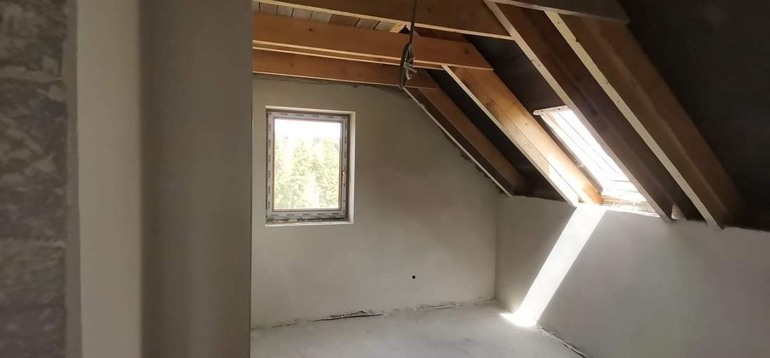 Poddasze z oknem dachowym i nowymi ścianami pokrytymi świeżym tynkiem, widoczne drewniane elementy konstrukcji dachu.