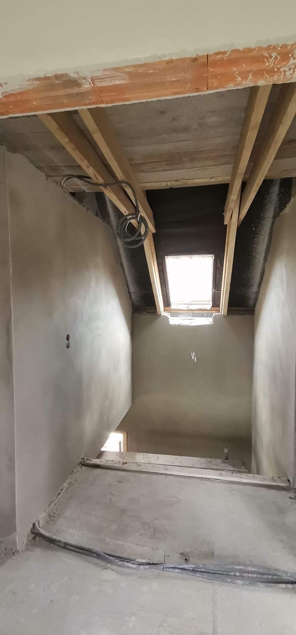 Wnętrze poddasza w trakcie prac wykończeniowych, widoczne surowe tynki na ścianach, drewniana konstrukcja dachu z oknem dachowym, oraz kable elektryczne zwisające z sufitu.