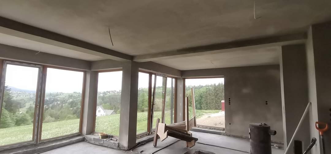 Wnętrze w stanie surowym z widokiem na krajobraz górski przez duże okna, ściany i sufit pokryte świeżym tynkiem, widoczne narzędzia i resztki materiałów budowlanych.