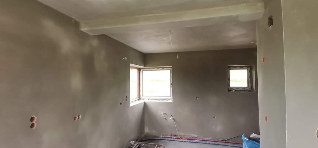 Surowe wnętrze po nałożeniu szarego tynku, widoczne okna z drewnianymi ramami, wystające przewody elektryczne i rury grzewcze przy podłodze.