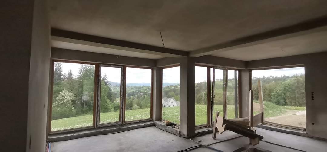 Surowy pokój z dużymi oknami i widokiem na zalesione wzgórza, ściany i sufit w szarym tynku, podłoga betonowa, widoczne niedoskonałości i zarysowania.