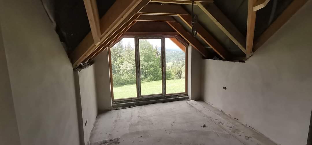 Poddasze z drewnianą konstrukcją dachu i oknem z widokiem na zieleń, ściany wstępnie przygotowane pod wykończenie, widoczne nierówności i ślady po pracach.