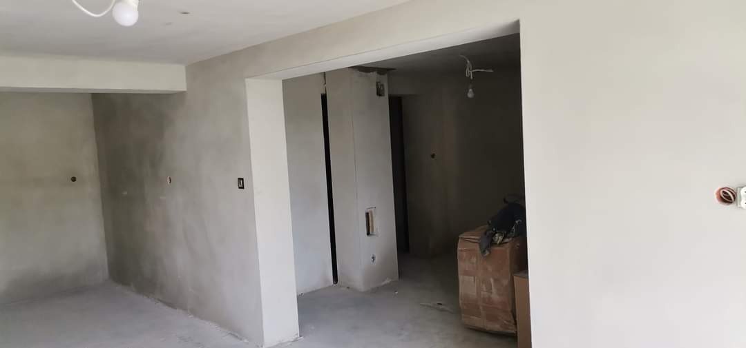 Wnętrze w trakcie prac wykończeniowych, ściany pokryte szarym tynkiem, widoczne puszki elektryczne i kartonowe pudła w przejściu do kolejnego pomieszczenia.