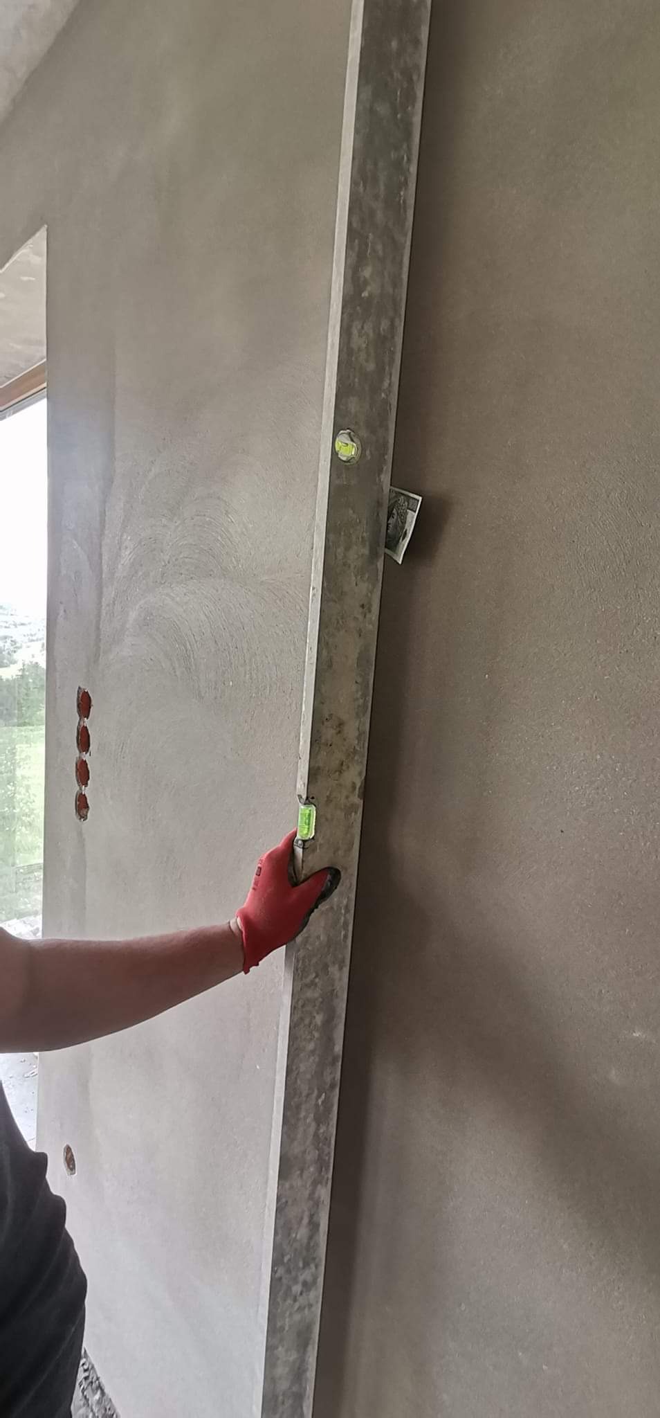 Sprawdzanie pionu ściany po nałożeniu warstwy wykończeniowej za pomocą poziomicy; widoczne czerwone rękawice ochronne i nieduży banknot włożony pod poziomicę.