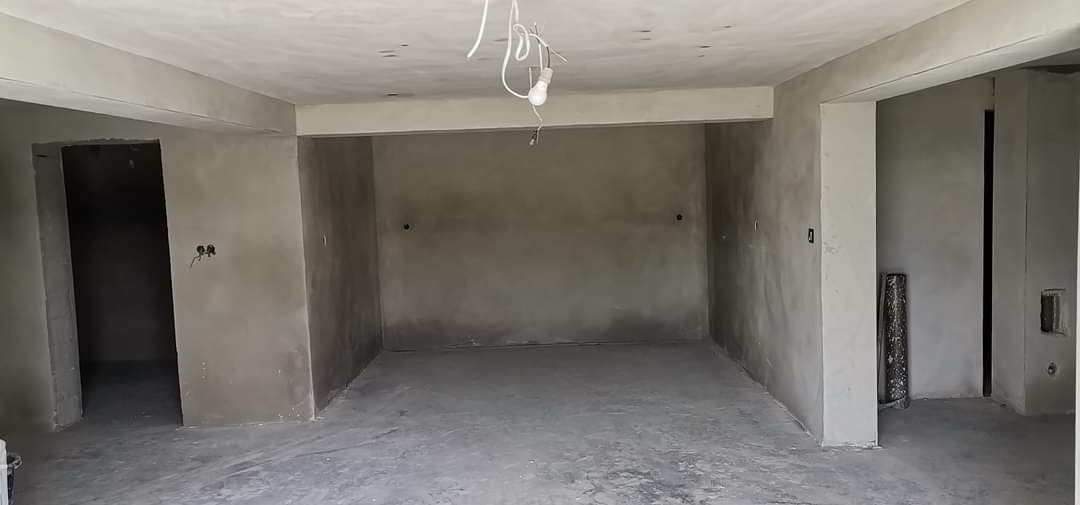 Surowe wnętrze po tynkowaniu maszynowym: szare ściany, widoczne kable oświetleniowe, otwarte przejścia do innych pomieszczeń, podłoga pokryta pyłem budowlanym.