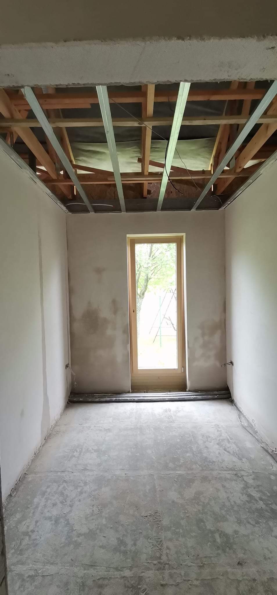 Surowy stan wnętrza pokoju przed wykończeniem, widoczne metalowe profile pod sufitem, wstępnie otynkowane ściany z widocznymi niedoskonałościami i surowa podłoga.