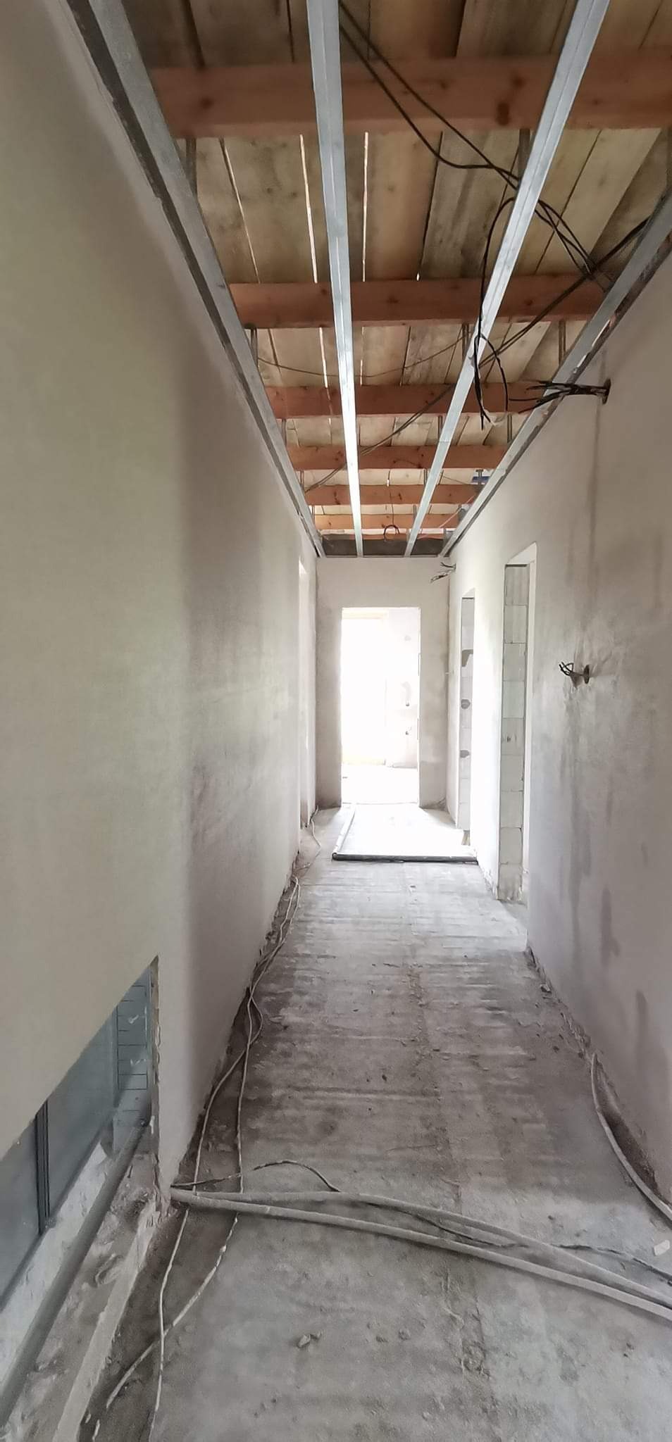 Korytarz w trakcie prac wykończeniowych: ściany pokryte świeżo nałożonym tynkiem, widoczna konstrukcja sufitu podwieszanego z metalowych profili i drewnianych łat, przewody elektryczne ułożone...