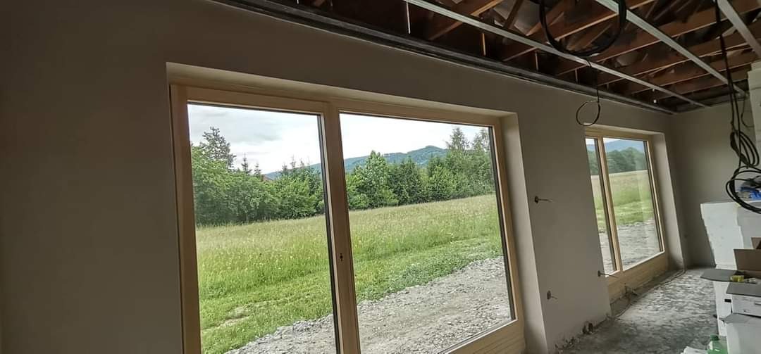 Wnętrze w trakcie prac wykończeniowych: ściany po nałożeniu tynku, duże okno z widokiem na łąkę i las, widoczne elementy konstrukcji sufitu podwieszanego.