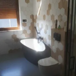 Nowoczesna łazienka z wolnostojącą wanną w kolorze antracytu, toaletą podwieszaną i heksagonalnymi płytkami imitującymi drewno na ścianie, oświetlona naturalnym światłem z okna z żaluzjami.