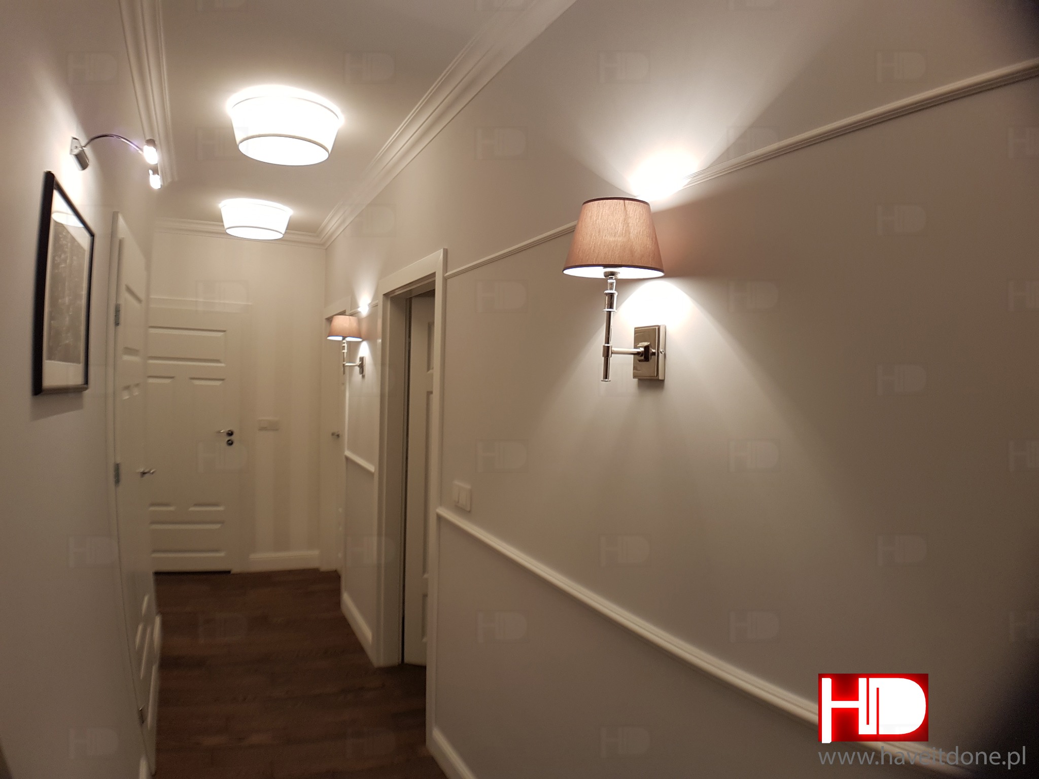 Korytarz w mieszkaniu w stylu klasycznym: drewniana podłoga, białe ściany z listwami, lampy ścienne z abażurami i sufitowe lampy wpuszczane.