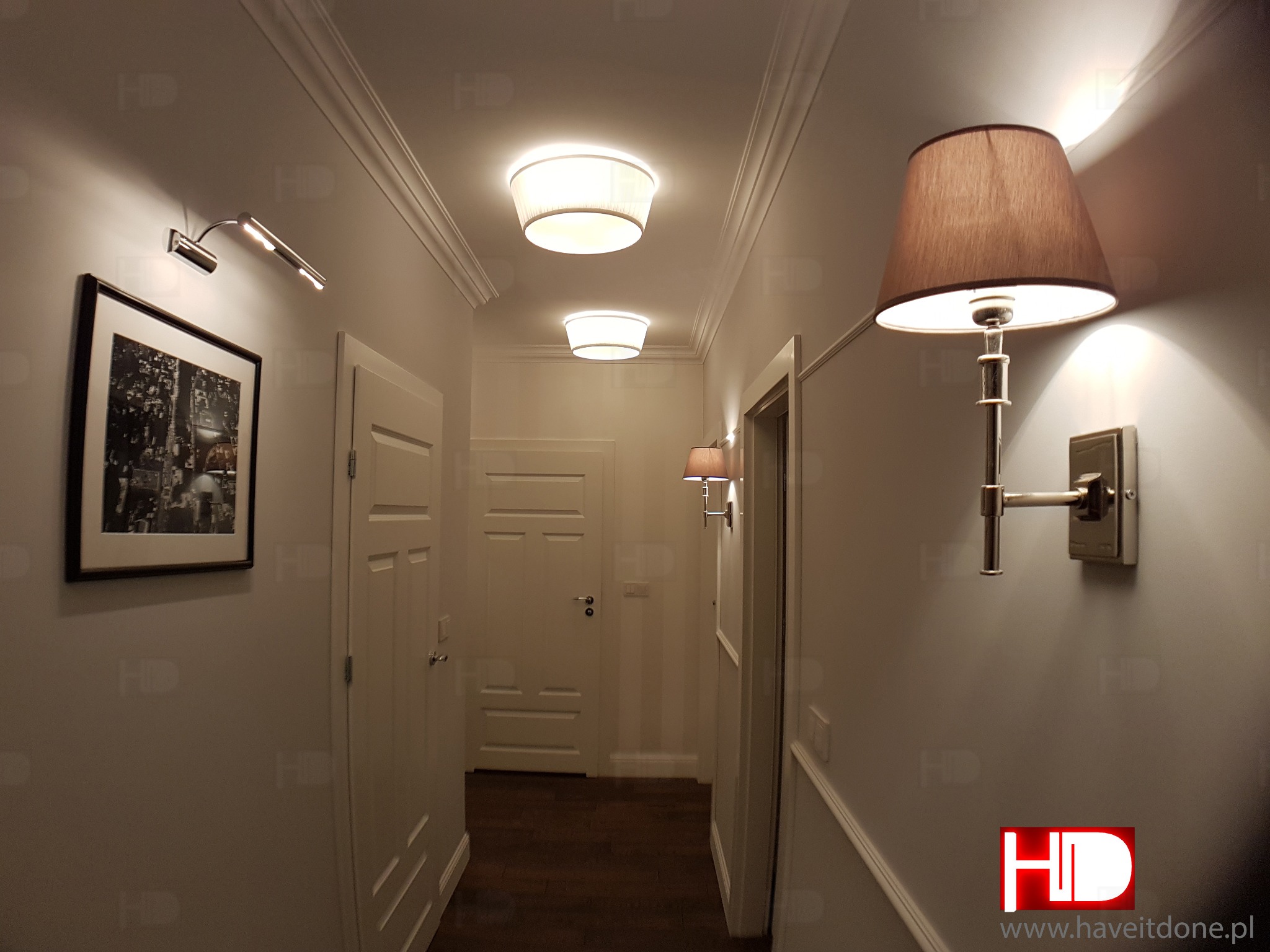 Korytarz w mieszkaniu z oświetleniem: lampy sufitowe i kinkiet z brązowym abażurem, na ścianie obraz w czarnej ramie.