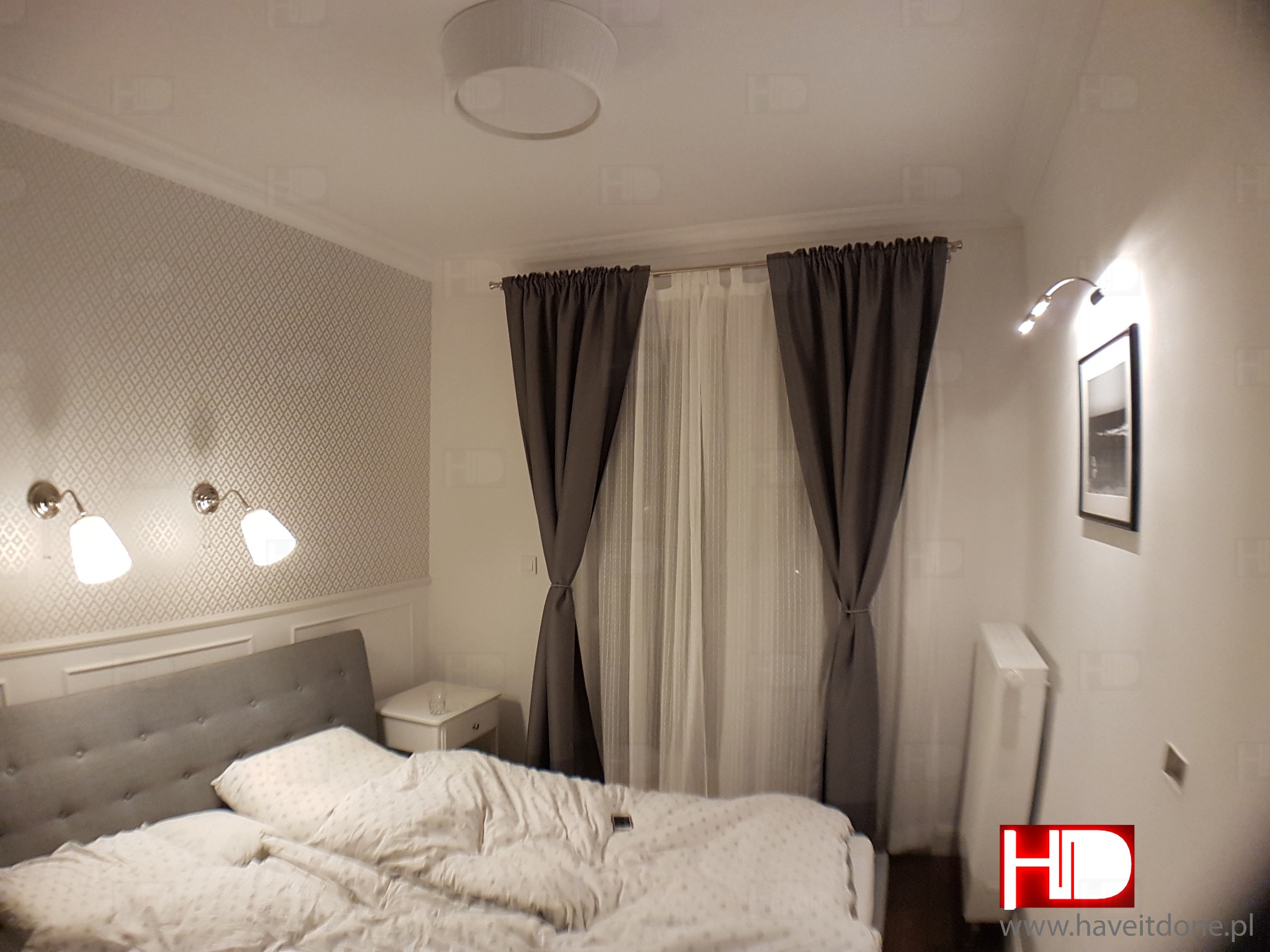 Sypialnia z szarą tapetą w romby, łóżkiem z szarym zagłówkiem, zasłonami w odcieniach szarości i białymi firanami, oświetlona lampami ściennymi i sufitową.