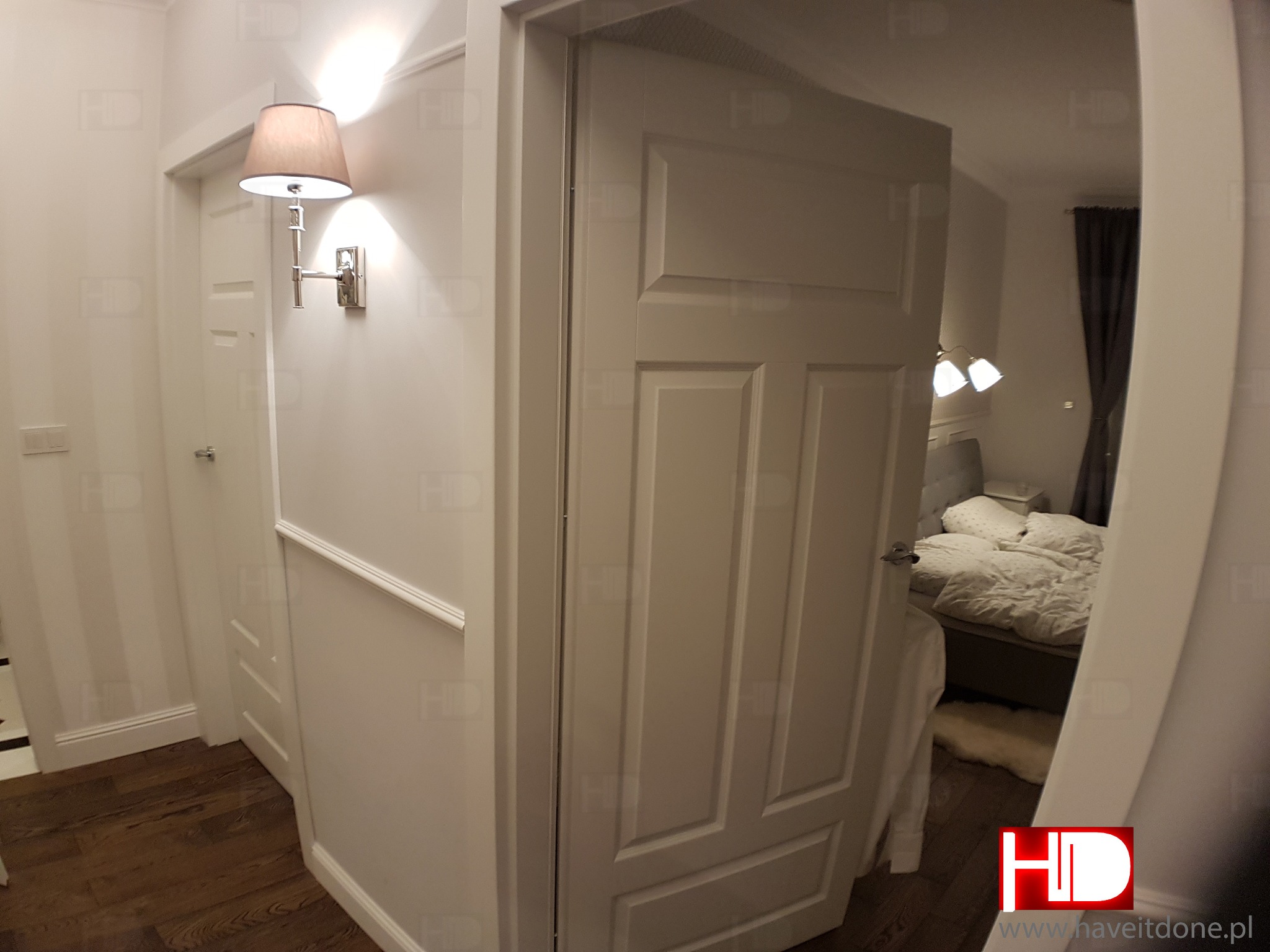 Widok na sypialnię z wejścia: białe ściany z dekoracyjną listwą, drewniana podłoga, otwarte białe drzwi, w tle sypialnia z łóżkiem i lampami