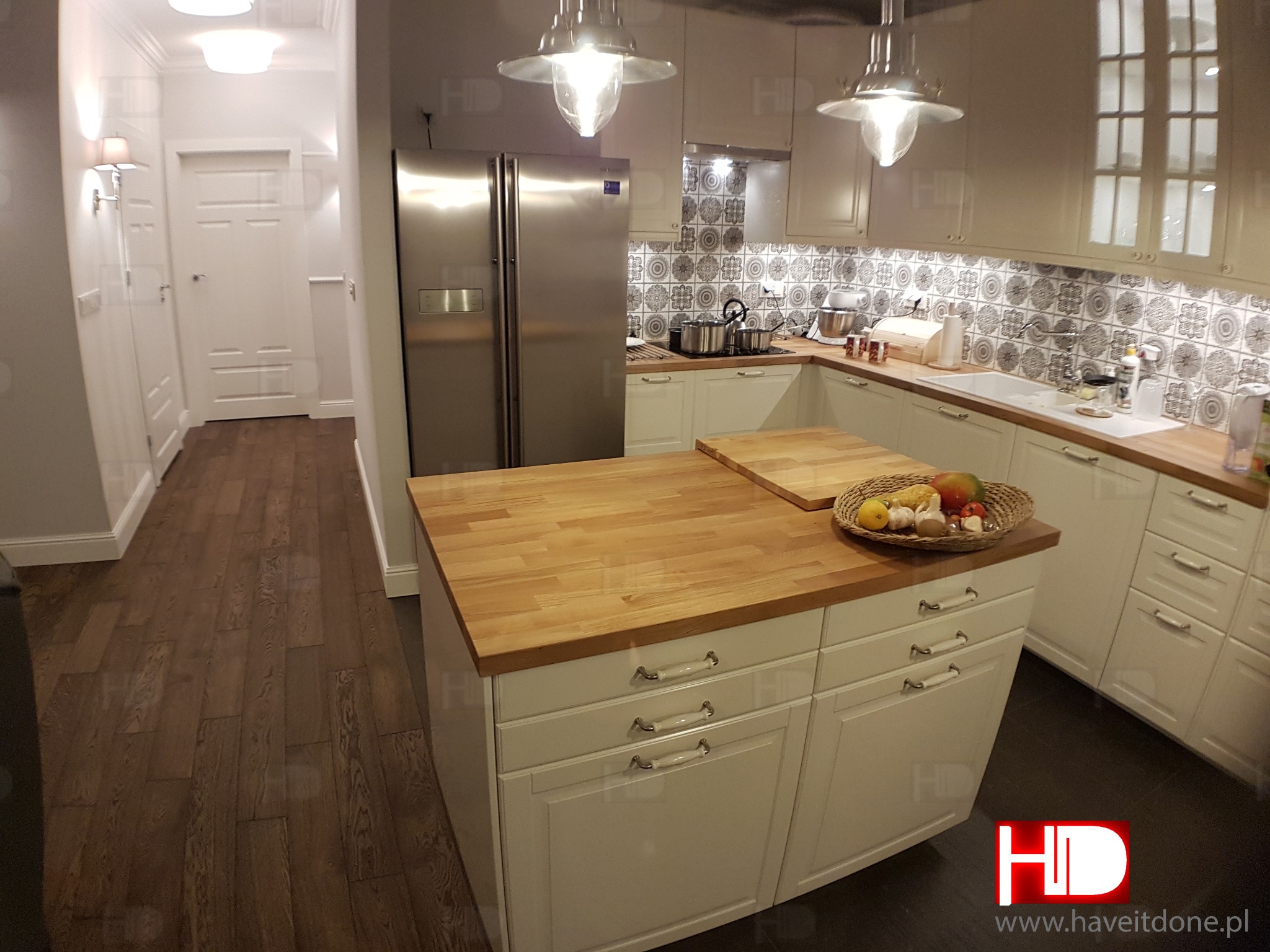 Jasna kuchnia w stylu klasycznym: białe szafki, drewniany blat wyspy z koszem owoców, stalowa lodówka i wzorzysta mozaika nad blatem roboczym.