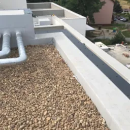 Płaski dach pokryty warstwą jasnych, otoczaków, z widocznymi elementami wentylacji i obróbkami blacharskimi, na tle zabudowy miejskiej i zieleni.