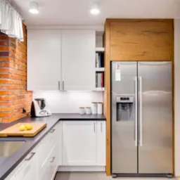 Jasna kuchnia z białymi szafkami, czarnym blatem i ceglaną ścianą przy oknie, wyposażona w dużą lodówkę z drewnianą obudową i zlew z baterią.