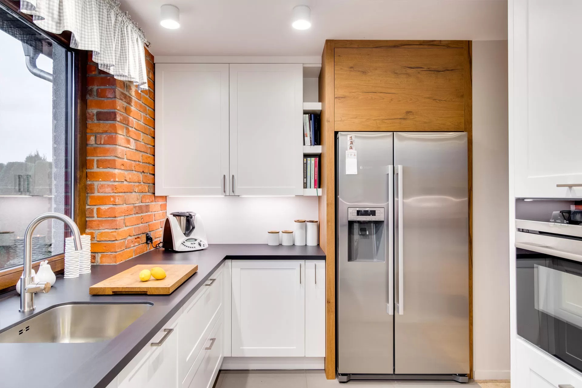 Jasna kuchnia z białymi szafkami, czarnym blatem i ceglaną ścianą przy oknie, wyposażona w dużą lodówkę z drewnianą obudową i zlew z baterią.