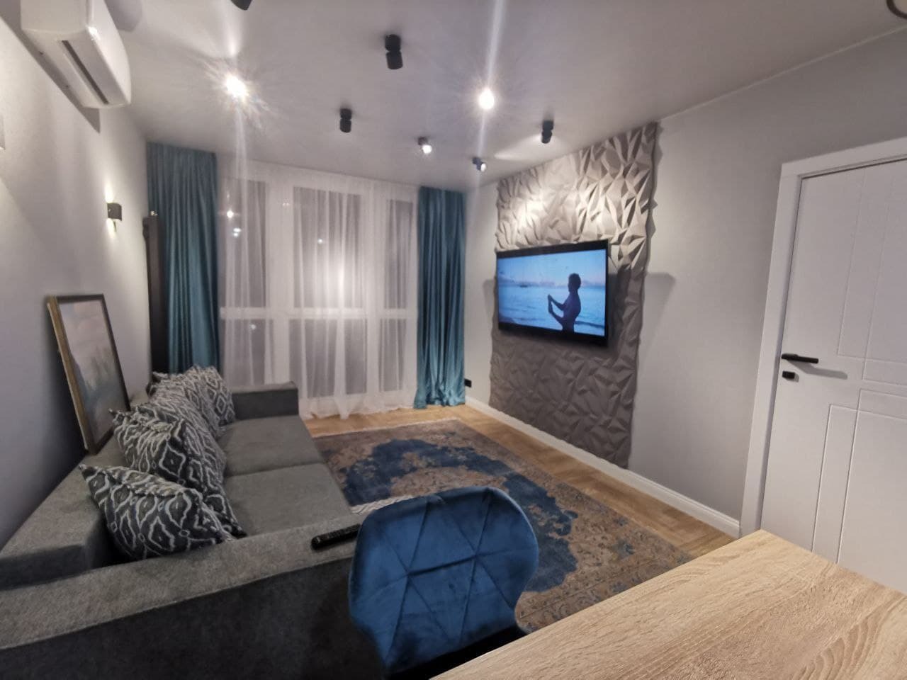 Nowoczesny salon z szarą sofą narożną, turkusowymi zasłonami i panelem 3D za telewizorem, oświetlony punktowo.