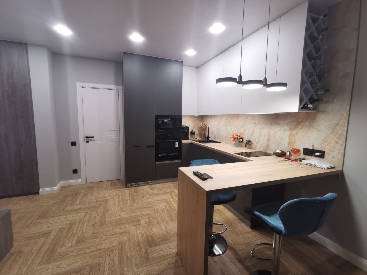 Nowoczesna kuchnia z wyspą i hokerami w odcieniach szarości, bieli i drewna, oświetlona trzema okrągłymi lampami wiszącymi, z parkietem ułożonym w jodełkę na podłodze.