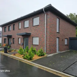 Nowoczesny segment bliźniaczy z czerwonej cegły, z szarymi roletami zewnętrznymi, małym ogródkiem z iglakami i podjazdem z ażurowych płyt betonowych w deszczowy dzień.