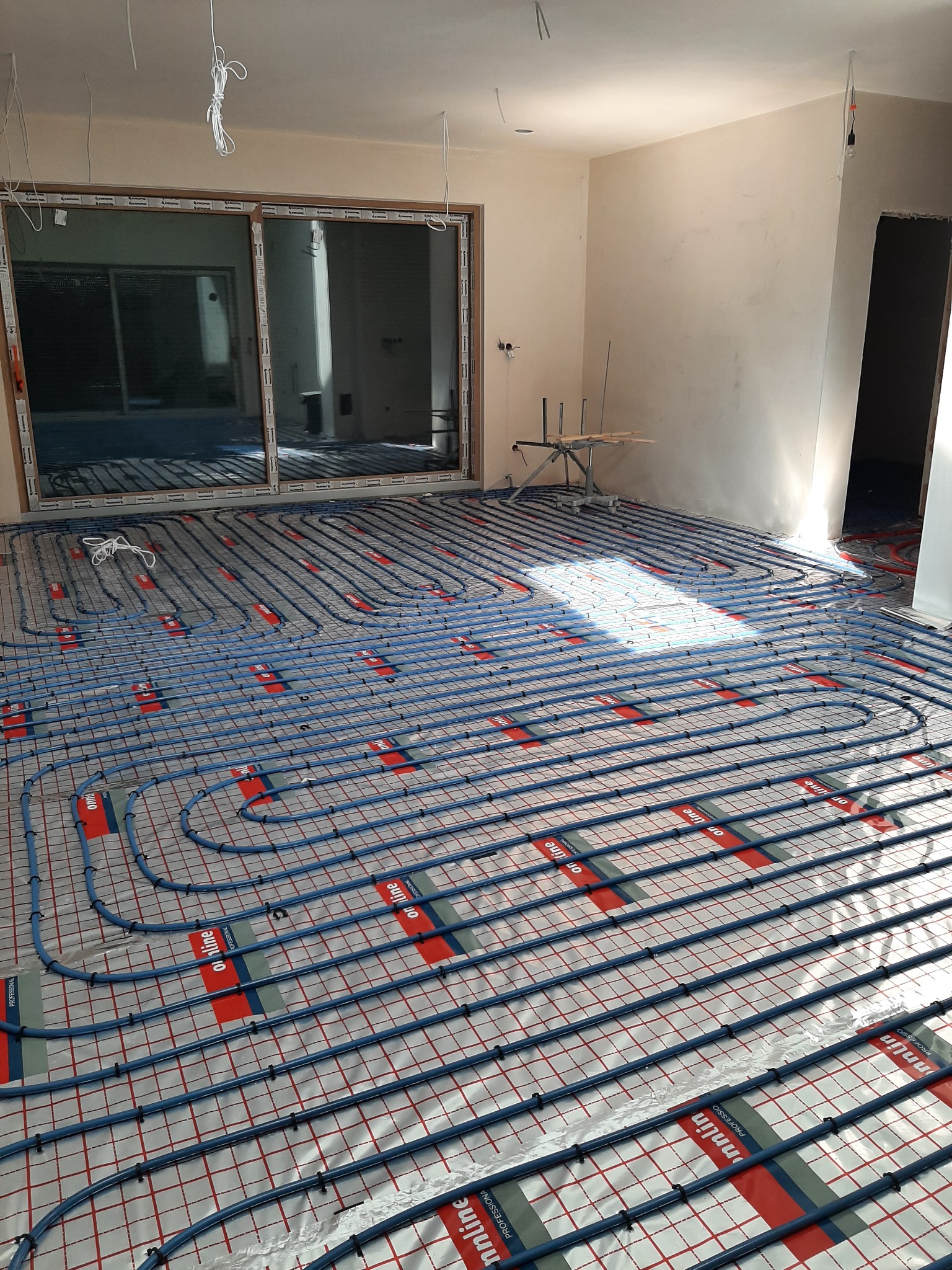 Rozłożona instalacja ogrzewania podłogowego z niebieskich rurek na folii z nadrukiem siatki i logo 'on line Professional', widoczne niedokończone ściany i duże okno przesuwne.