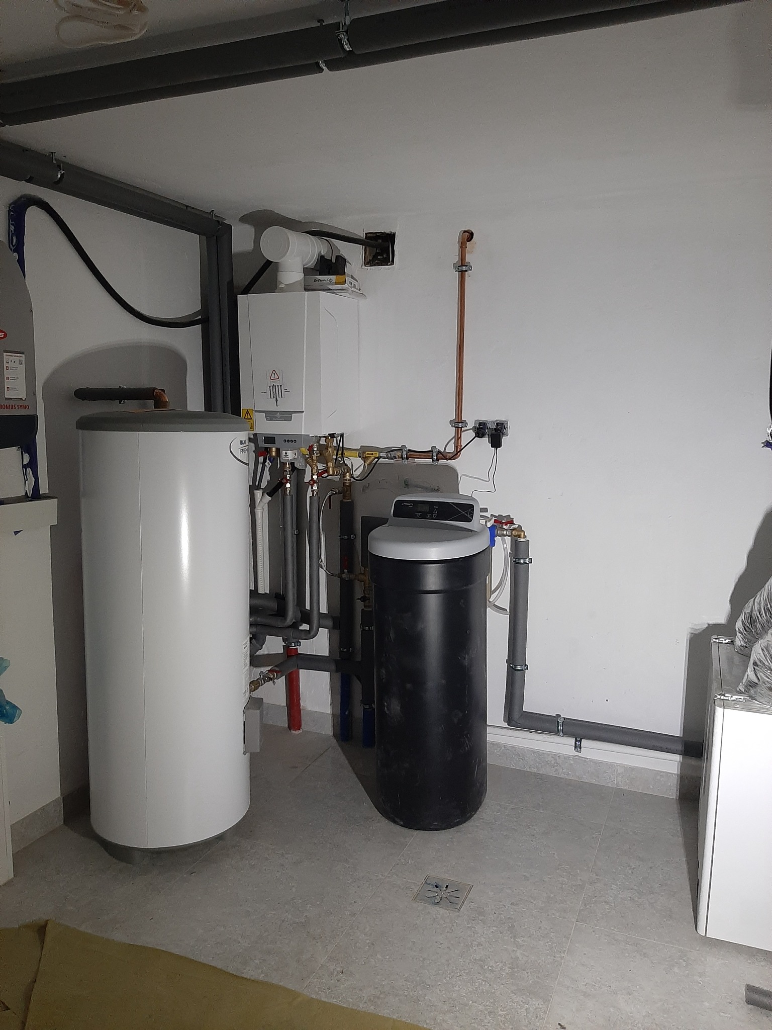 Kotłownia z białym zasobnikiem ciepłej wody, piecem gazowym i czarnym zmiękczaczem wody, widoczne rury miedziane i szare instalacyjne, oświetlenie sufitowe.