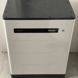 LONGTERM SPÓŁKA Z OGRANICZONĄ ODPOWIEDZIALNOŚCIĄ - Biało-czarny magazyn energii GoodWe z widocznym wskaźnikiem naładowania umieszczony w rogu pomieszczenia.