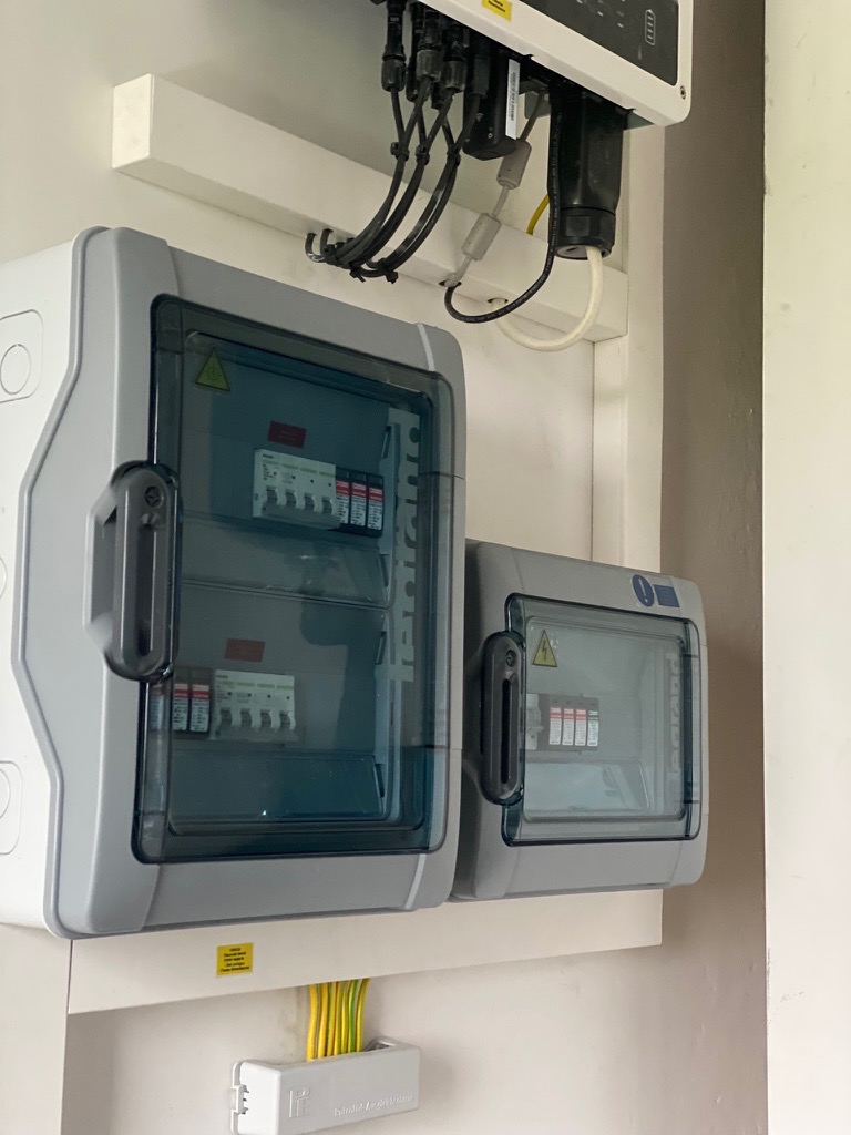 Dwa szare rozdzielnice elektryczne z przezroczystymi drzwiczkami, zamontowane na białej ścianie, z widocznymi kablami i zabezpieczeniami wewnątrz.