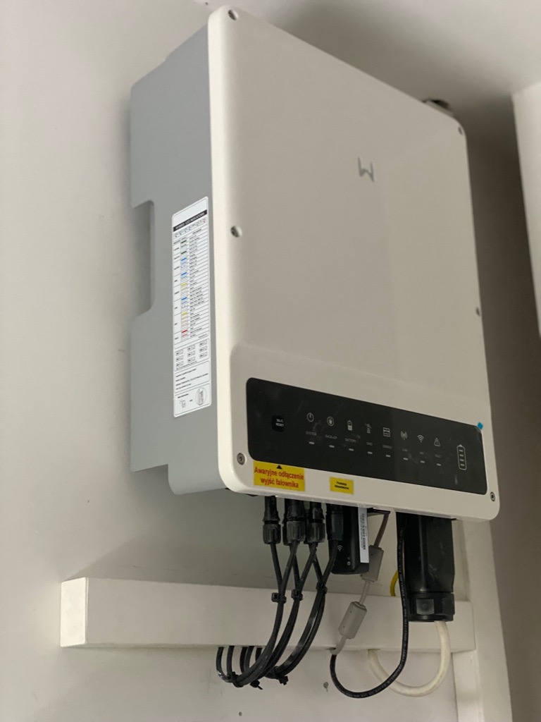 Biały falownik z widocznymi przewodami i żółtą naklejką ostrzegawczą, zamontowany na ścianie na białej półce.