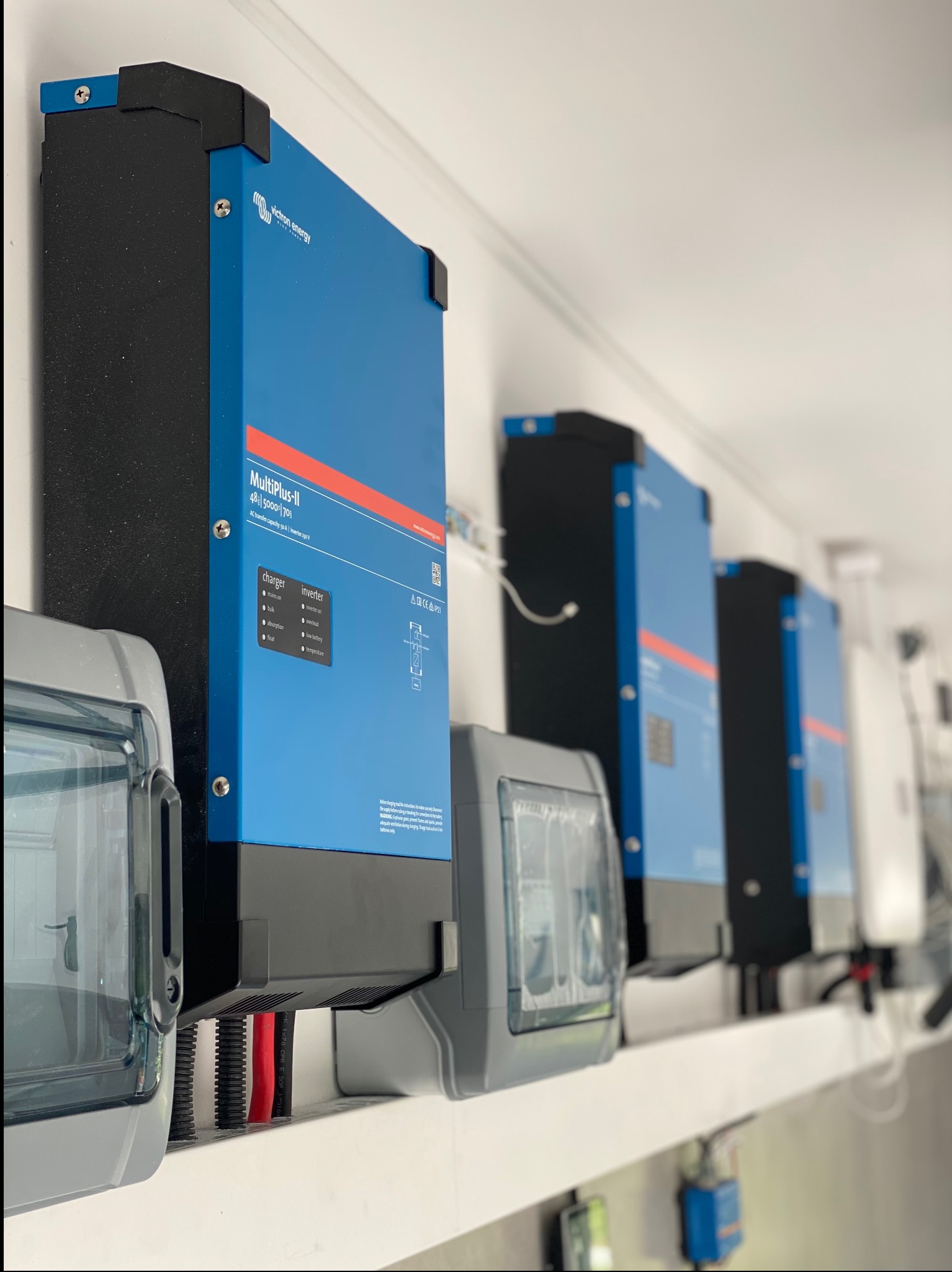 Rząd niebieskich inwerterów Victron Energy MultiPlus-II zamontowanych na białej ścianie, z widocznymi przewodami i szarą skrzynką zabezpieczającą po lewej stronie.