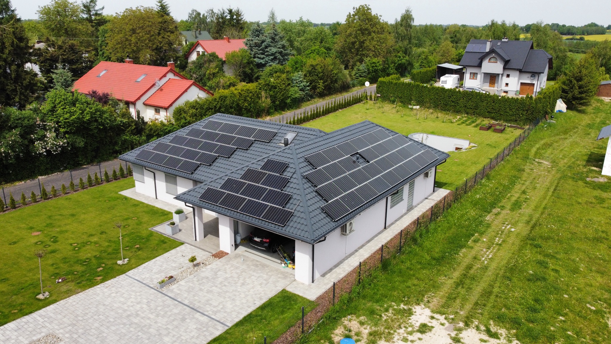 Widok z drona na dom jednorodzinny z szarym dachem pokrytym panelami słonecznymi, garaż z samochodem, zadbany trawnik i kostka brukowa na podjeździe.