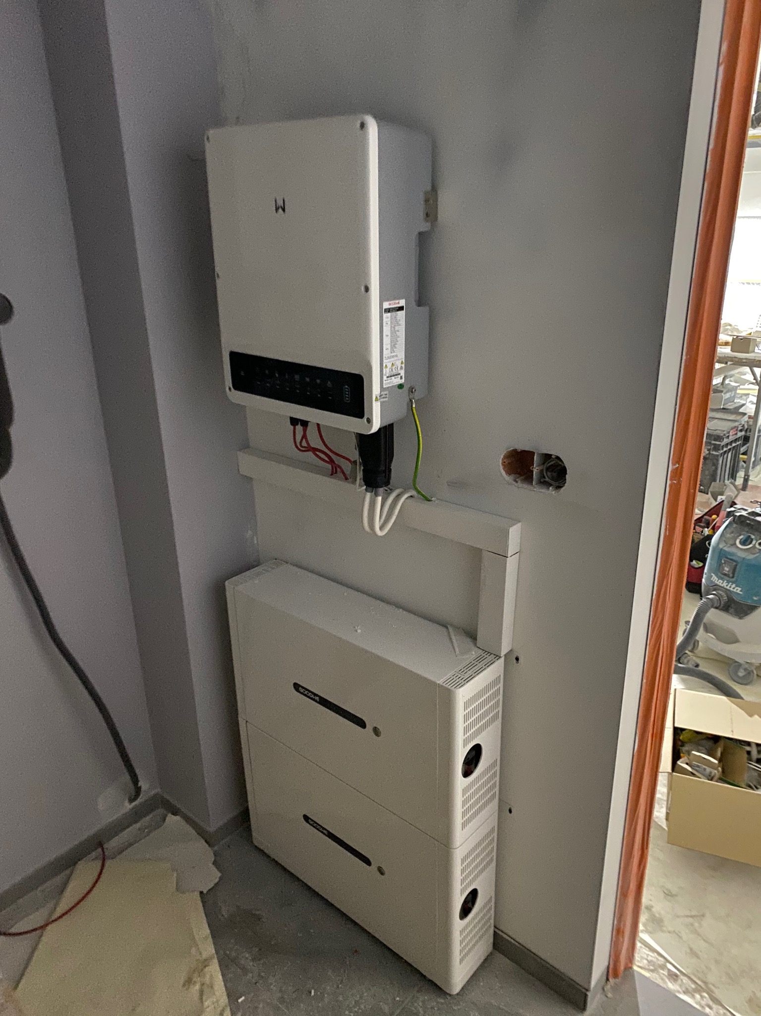 Biały inwerter z modułem bateryjnym na szarej ścianie w trakcie montażu, widoczne okablowanie i otwór w ścianie na przewody, w tle narzędzia i materiały budowlane.