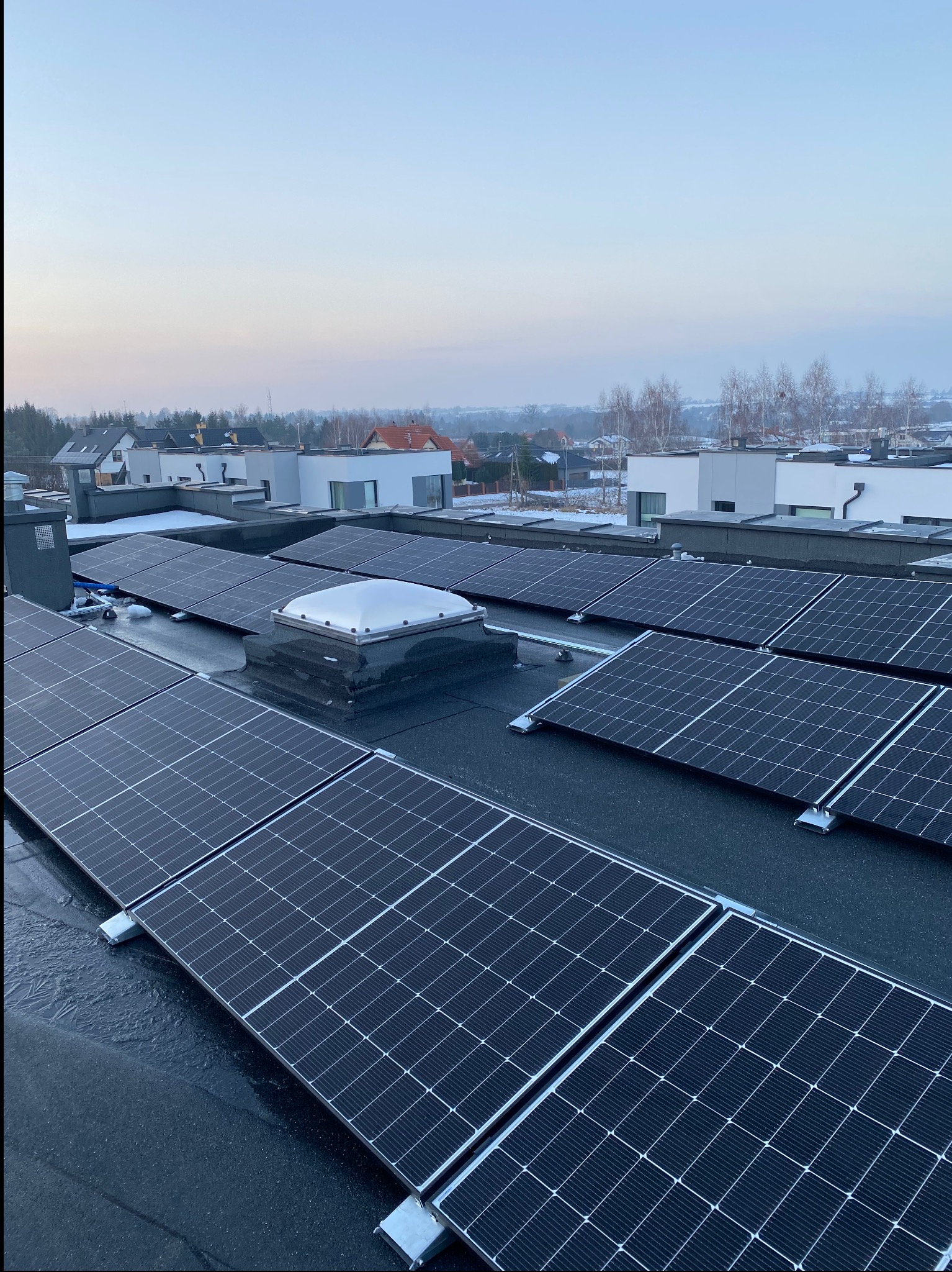 Panele słoneczne na płaskim dachu z widocznym świetlikiem, w tle zabudowa jednorodzinna i lekka mgła.