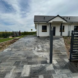 KARO SPÓŁKA Z OGRANICZONĄ ODPOWIEDZIALNOŚCIĄ - Ułożona kostka brukowa w odcieniach szarości prowadząca do nowoczesnego domu z ciemną dachówką, widoczny fragment metalowej bramy i osoba pracująca w tle przy ogrodzeniu.