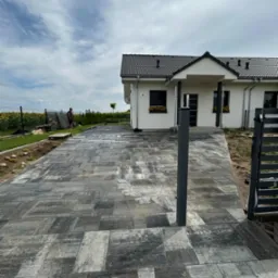 Ułożona kostka brukowa w odcieniach szarości prowadząca do nowoczesnego domu z ciemną dachówką, widoczny fragment metalowej bramy i osoba pracująca w tle przy ogrodzeniu.