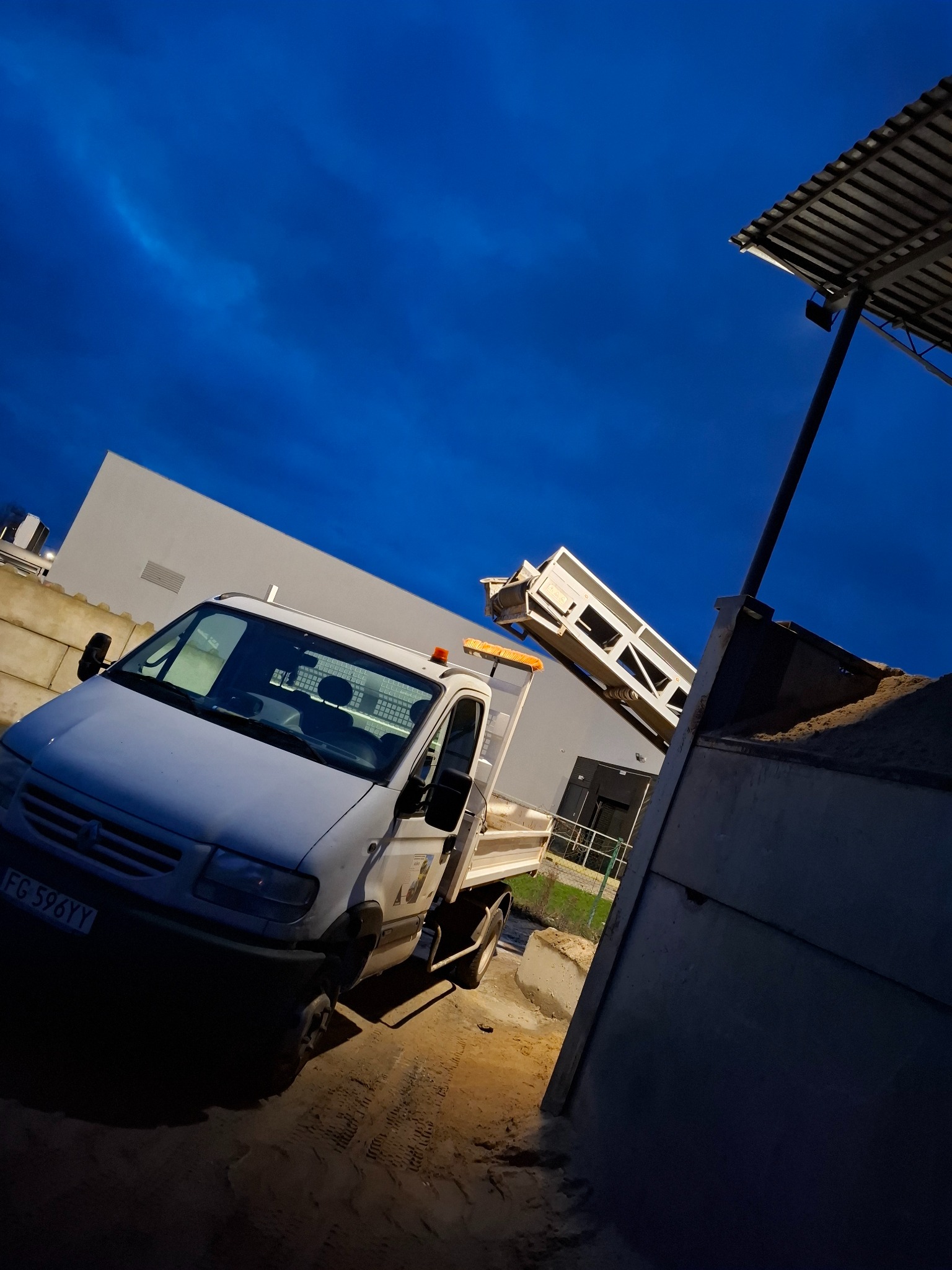 Biała wywrotka z podniesioną skrzynią ładunkową, wysypująca ziemię na placu budowy pod błękitnym niebem.