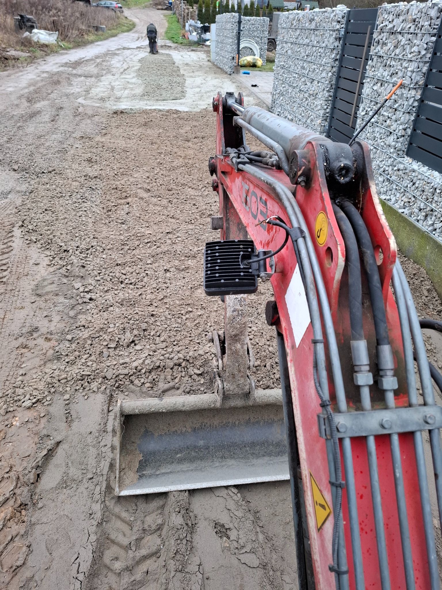 Czerwony minikoparka z łyżką w trakcie prac ziemnych, widoczne świeżo rozplantowane kruszywo na podjeździe i osoba wyrównująca teren w oddali. Gabionowe ogrodzenie w tle.