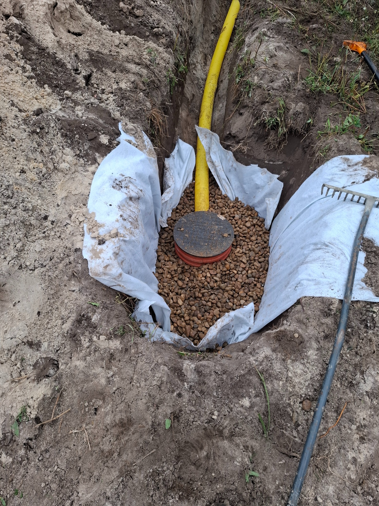 Wykop w ziemi wyłożony geowłókniną, wypełniony drobnymi kamieniami, z żółtą rurą i czarną pokrywą rewizyjną w środku, obok leży grabie.